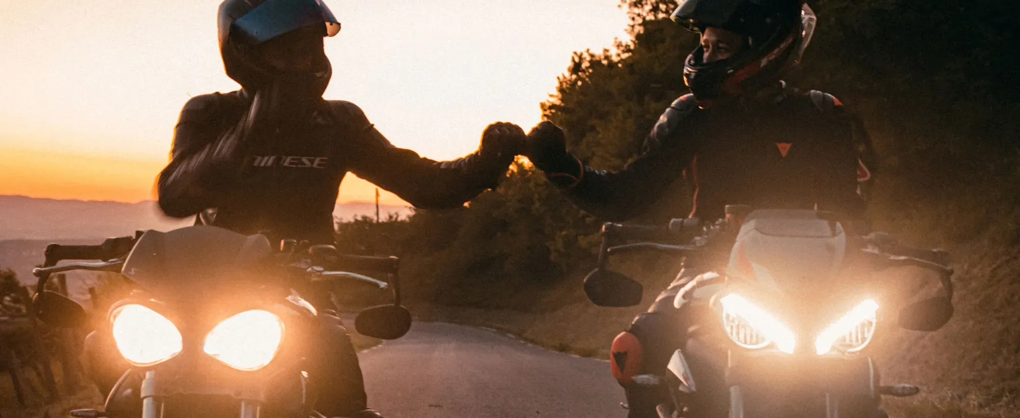 Deux motards se saluent, moteurs tournants, sur une route de col au coucher du soleil